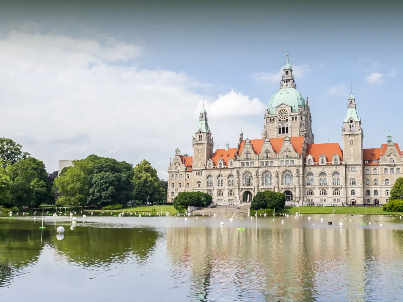 Das Neue Rathaus in Hannover mit seiner markanten grünen Kuppel, das an einem See liegt und sich leicht im Wasser spiegelt. Es ist von vielen grünen Bäumen umgeben.