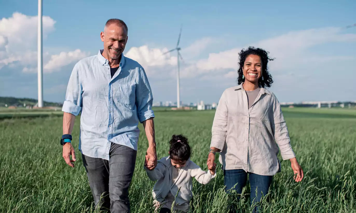Eine Familie aus Mann, Frau und Kind wandert Hand in Hand über ein Feld. Im Hintergrund ist ein Windrad zu sehen.