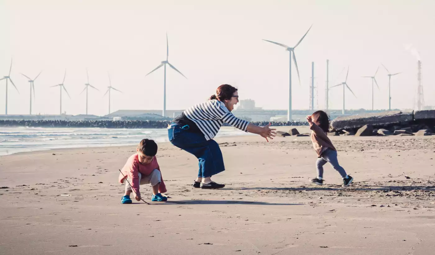 Eine Frau und zwei Kinder spielen am Strand. Im Hintergrund ist ein Windpark zu sehen.