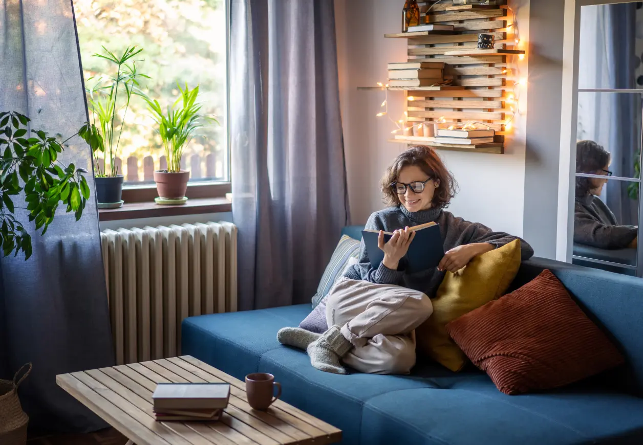 Eine Frau liest ein Buch in einem gemütlichen Wohnzimmer, im Hintergrund ist ein Heizkörper zu sehen.