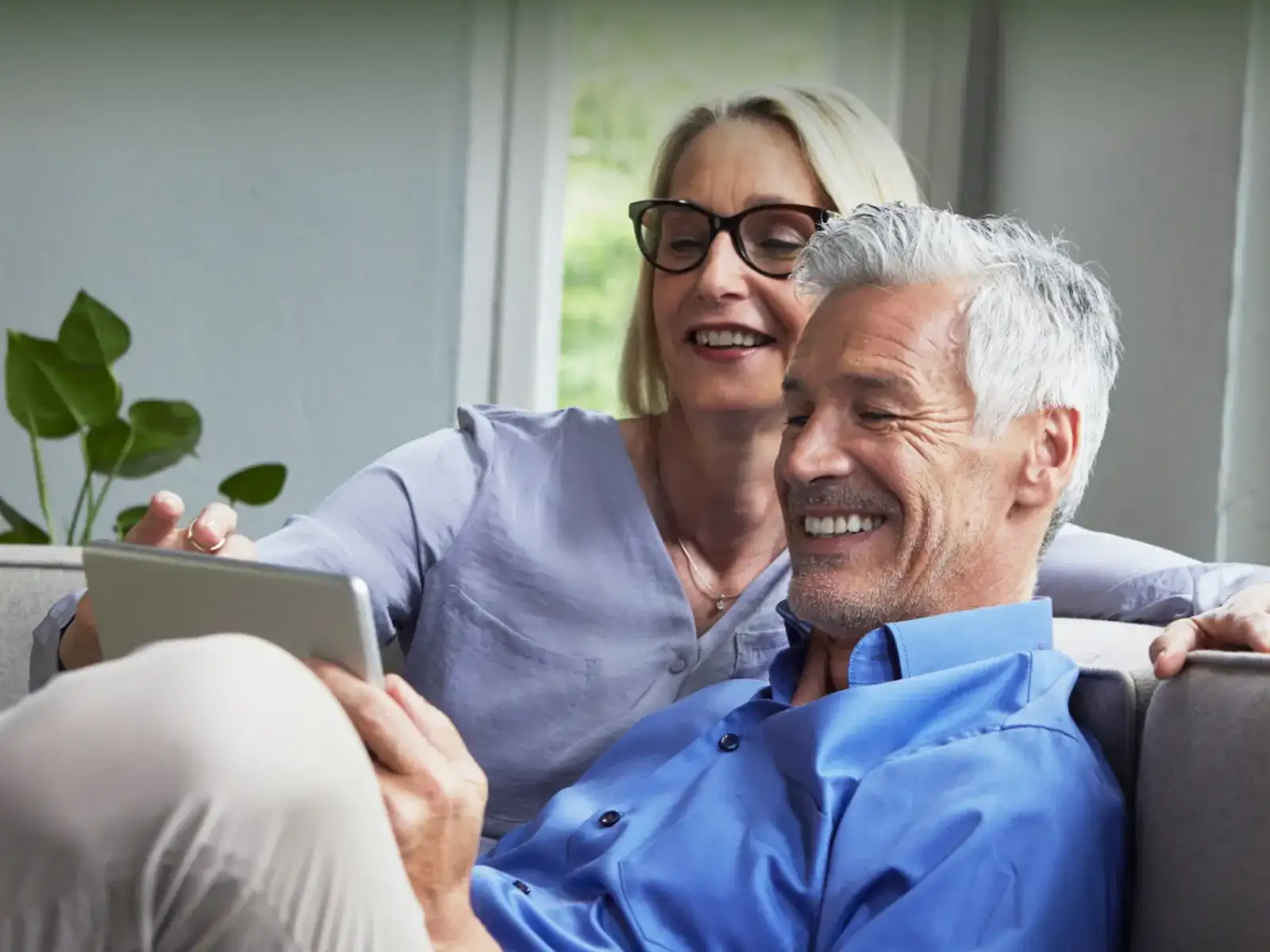Älteres, lächelndes Paar sitzt auf einem Sofa und schaut gemeinsam auf ein Tablet.