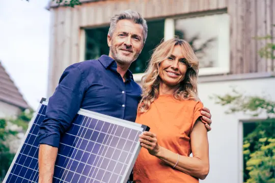 Paar steht mit einem Solarpanel vorm Haus.