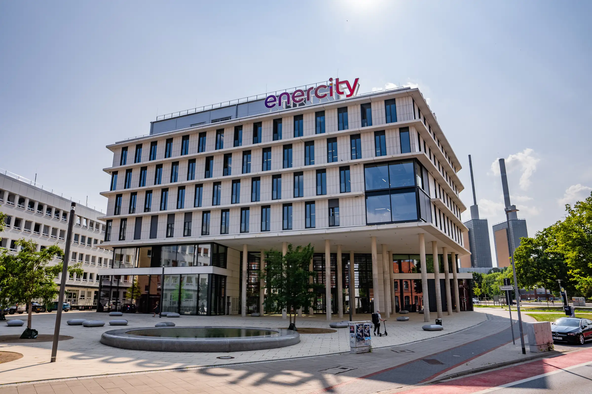 Moderne enercity-Konzernzentrale in einem hellen Bürogebäude mit großem „enercity“-Logo auf dem Dach, davor ein Platz mit Brunnen und Bäumen.