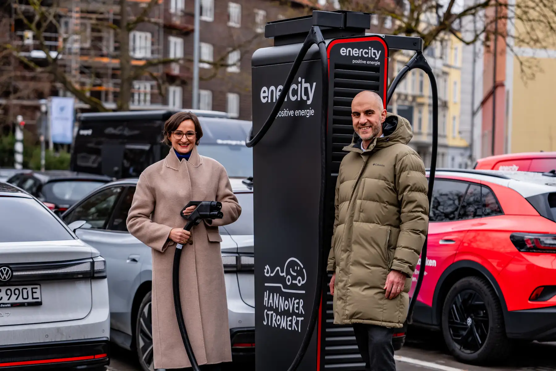 enercity-CEO Aurélie Alemany und Hannovers Oberbürgermeister Belit Onay stehen lächelnd neben einer enercity-Ladesäule für E-Autos. Alemany hält einen Ladestecker in der Hand, im Hintergrund sind geparkte Fahrzeuge auf einer Straße zu sehen.