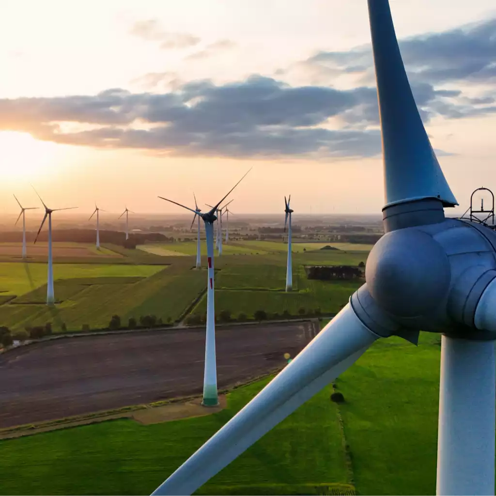 Windpark mit mehreren Windrädern über grünen Feldern bei Sonnenuntergang, ein Rotorblatt im Vordergrund.